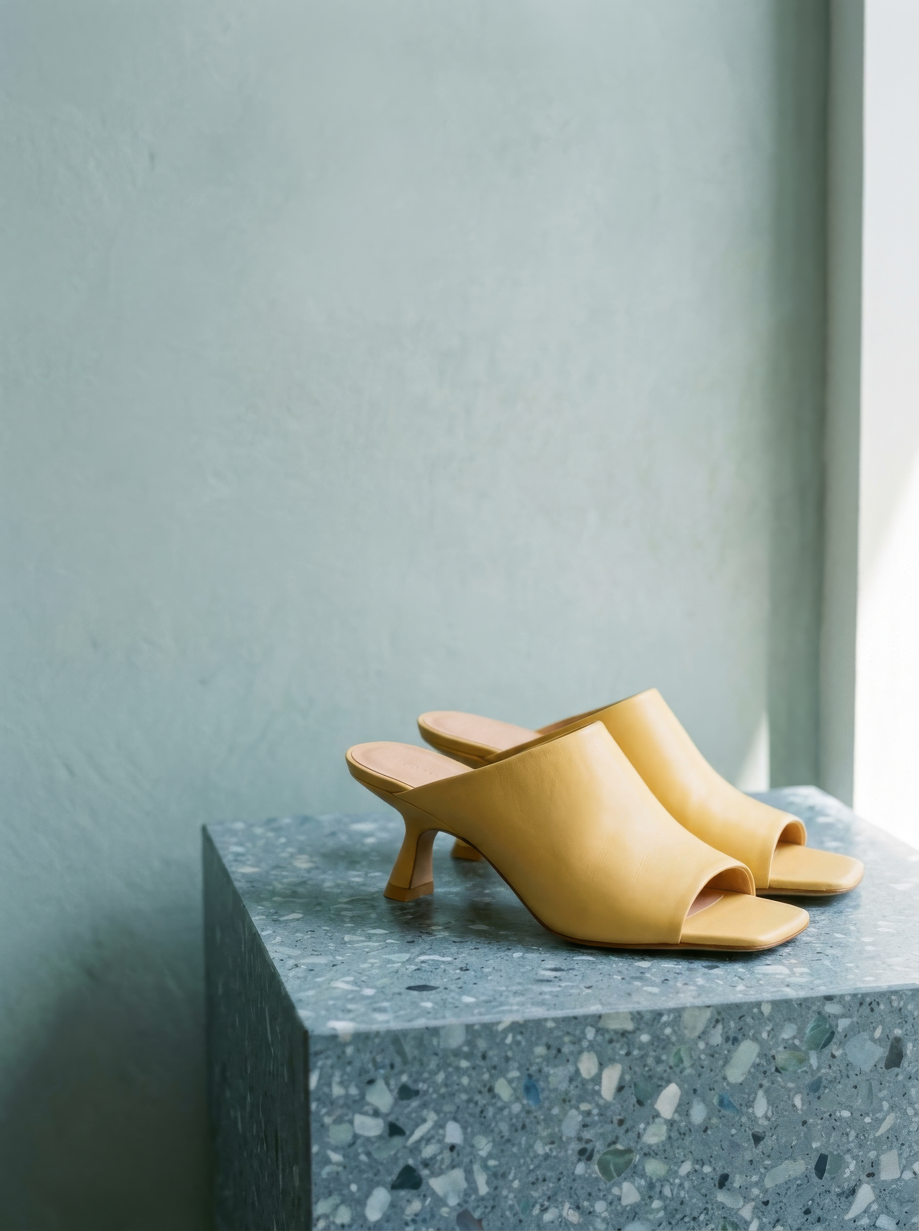 KAMI butter-yellow leather heeled mules with a square open toe, displayed on a blue terrazzo plinth against a soft sage wall.