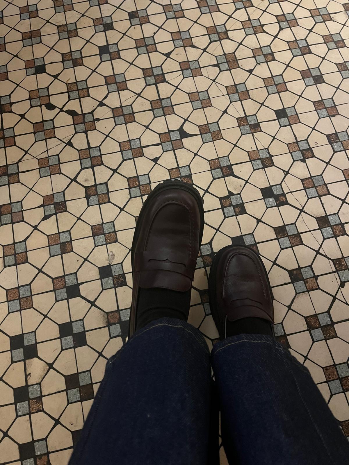 Person wearing oxblood loafers and blue jeans standing on a patterned tile floor.