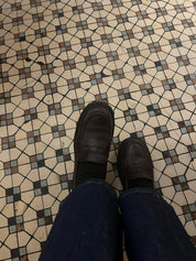 Person wearing oxblood loafers and blue jeans standing on a patterned tile floor.