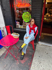 Woman sitting on a red chair in front of a store with a neon sign wearing yellow socks and black loafers.