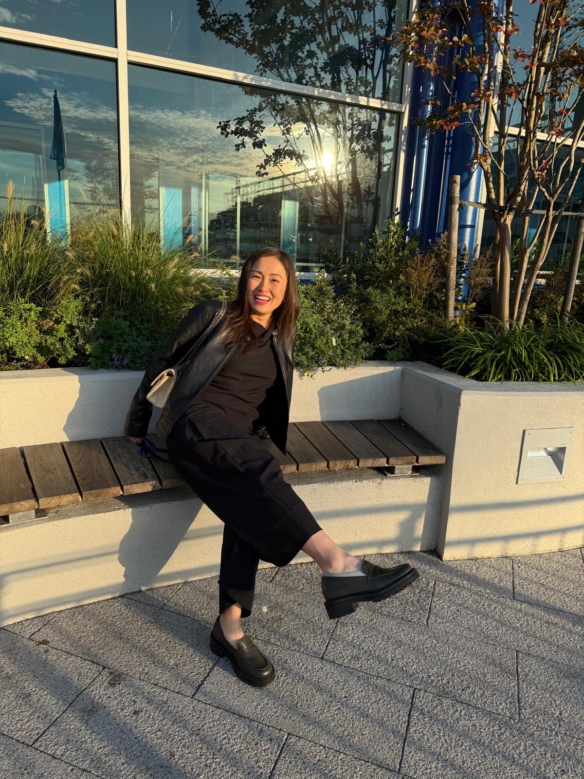 Woman sitting on a bench outdoors with plants and building reflections in the background wearing green chunky leather loafers.