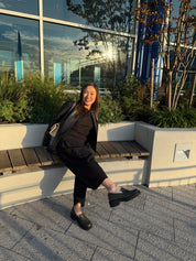 Woman sitting on a bench outdoors with plants and building reflections in the background wearing green chunky leather loafers.